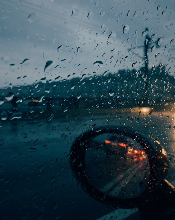 Rainy weather. Raindsrops outside car with auto mirror, unrecognizable driver drives car at bad weather during evening. Vertical shot. Weather conditions and transportation