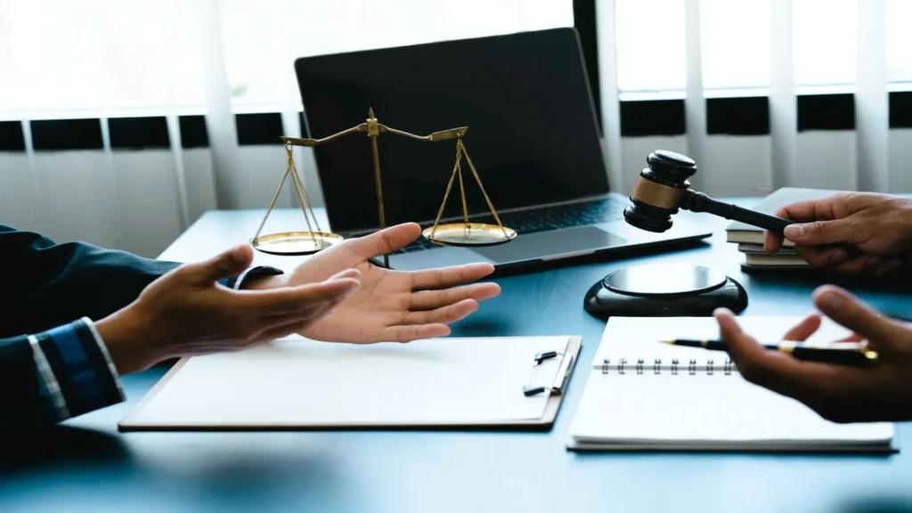 Lawyers discussing a case with scales of justice, gavel, notebook, and pen on a desk with a laptop in the background.